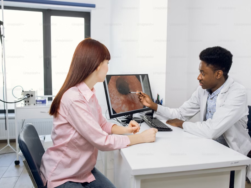 
A imagem mostra um consultório médico com uma médica e um paciente sentados um de frente para o outro, ambos olhando para a tela de um computador. 