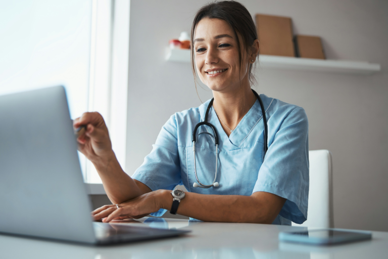 A imagem mostra uma profissional da área da saúde, possivelmente uma médica ou enfermeira, vestindo um jaleco azul claro e com um estetoscópio pendurado no pescoço. Ela está sentada em uma mesa, sorrindo e interagindo com um laptop — aparentemente durante uma videoconferência, consulta online ou atividade administrativa.