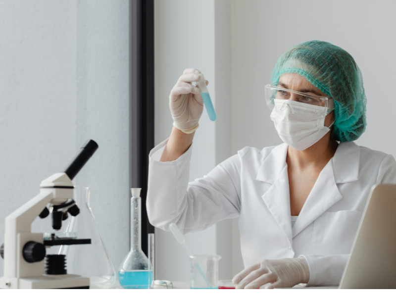 A imagem mostra uma pesquisadora ou profissional de saúde em um ambiente de laboratório, envolvida em atividades de análise científica. Ela está usando equipamentos de proteção individual (EPI)