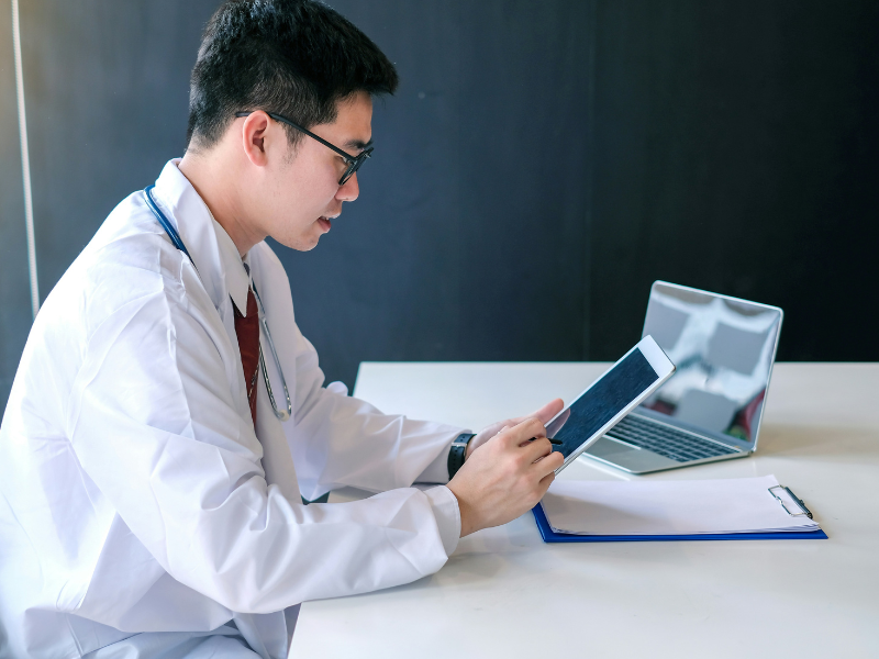 A imagem mostra um médico jovem sentado à mesa em um ambiente de trabalho moderno. Ele está vestindo um jaleco branco, com um estetoscópio pendurado no pescoço, o que indica sua atuação na área da saúde.