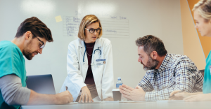 Médica de jaleco branco conversa com um homem sentado em uma mesa de escritório.
