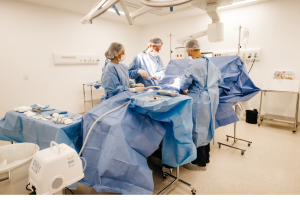 A imagem mostra uma equipe médica realizando uma cirurgia em um centro cirúrgico moderno. Todos os profissionais estão usando roupas estéreis, como aventais cirúrgicos azuis, luvas, máscaras e toucas, que são padrões de segurança em ambientes hospitalares para prevenir infecções.