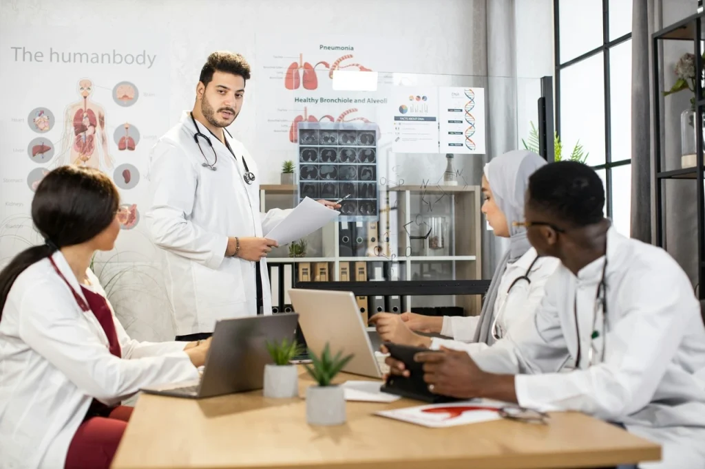 Grupo de médicos reunidos em uma sala de conferência moderna, discutindo temas relacionados à administração e negócios hospitalares. Eles estão em trajes clínicos — jalecos brancos e estetoscópios —, alguns com tablets e notebooks, analisando gráficos e documentos. Ao fundo, um quadro digital exibe indicadores de gestão hospitalar, como fluxo de pacientes, orçamento e desempenho. A cena transmite colaboração, planejamento estratégico e integração entre a prática médica e a gestão hospitalar