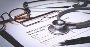 Estetoscópio cinza e óculos sobre prancheta com formulário de histórico médico.