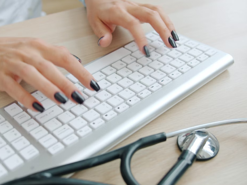 A imagem é um close-up que foca nas mãos de uma pessoa digitando em um teclado de computador.

As unhas da pessoa estão pintadas com esmalte preto. Em primeiro plano, no canto inferior direito, há um estetoscópio preto, indicando que a pessoa é uma profissional de saúde.