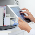 A imagem foca nas mãos de uma pessoa (provavelmente um profissional de saúde, como um médico) interagindo com um tablet digital.