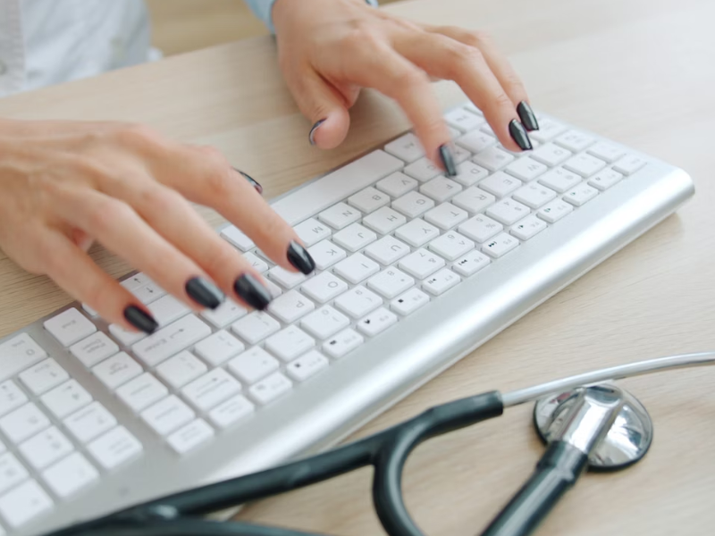 A imagem mostra as mãos de uma pessoa digitando em um teclado branco, provavelmente em um ambiente médico, já que há um estetoscópio sobre a mesa ao lado.