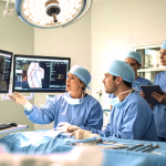 Equipe médica em centro cirúrgico analisa exames anatômicos em múltiplos monitores.