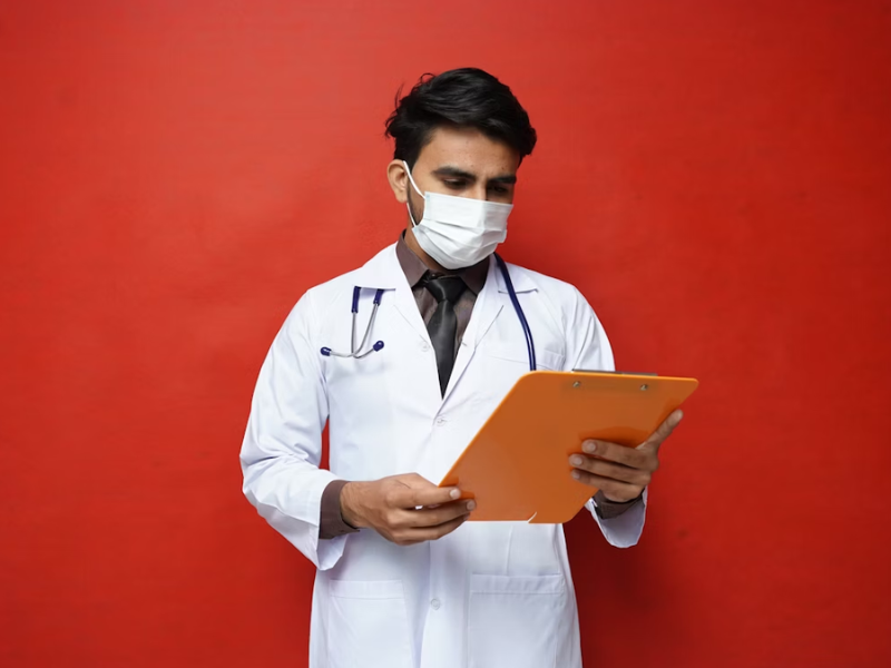 A imagem mostra um profissional de saúde em pé, vestindo um jaleco branco, camisa escura e gravata, além de uma máscara cirúrgica.