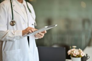 A imagem mostra um profissional da área da saúde usando um tablet. A pessoa veste um jaleco branco e tem um estetoscópio pendurado no pescoço, indicando que pode ser um médico ou outro profissional clínico.