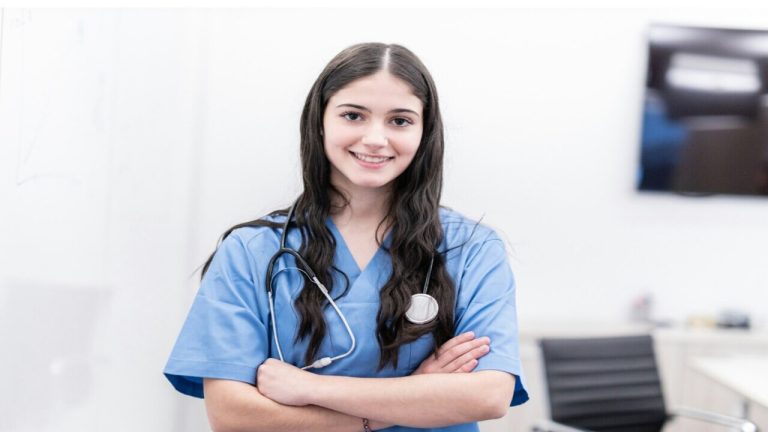 A imagem mostra uma profissional de saúde jovem sorrindo, vestindo uniforme azul e com um estetoscópio pendurado no pescoço. Ela está de braços cruzados e parece confiante.