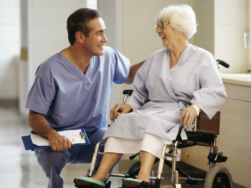A foto retrata uma interação calorosa e profissional entre um profissional de saúde (provavelmente um enfermeiro ou cuidador) e uma paciente idosa em um ambiente que parece ser um corredor ou quarto de hospital/clínica.