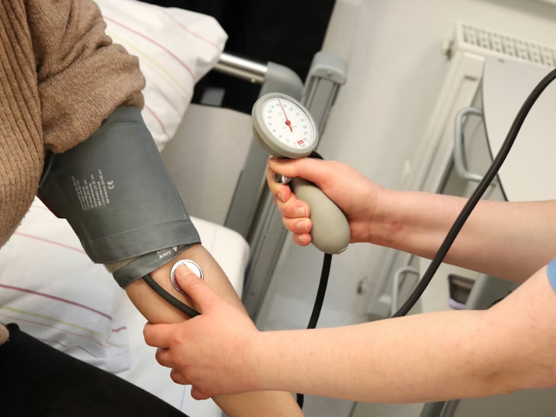 A foto é um close-up focado na ação de um profissional de saúde verificando a pressão arterial de um paciente, destacando um momento essencial do cuidado clínico.