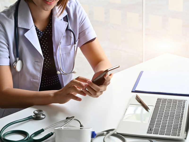 Médica ou profissional da saúde usando smartphone em ambiente clínico, com estetoscópio, laptop e equipamento médico na mesa.