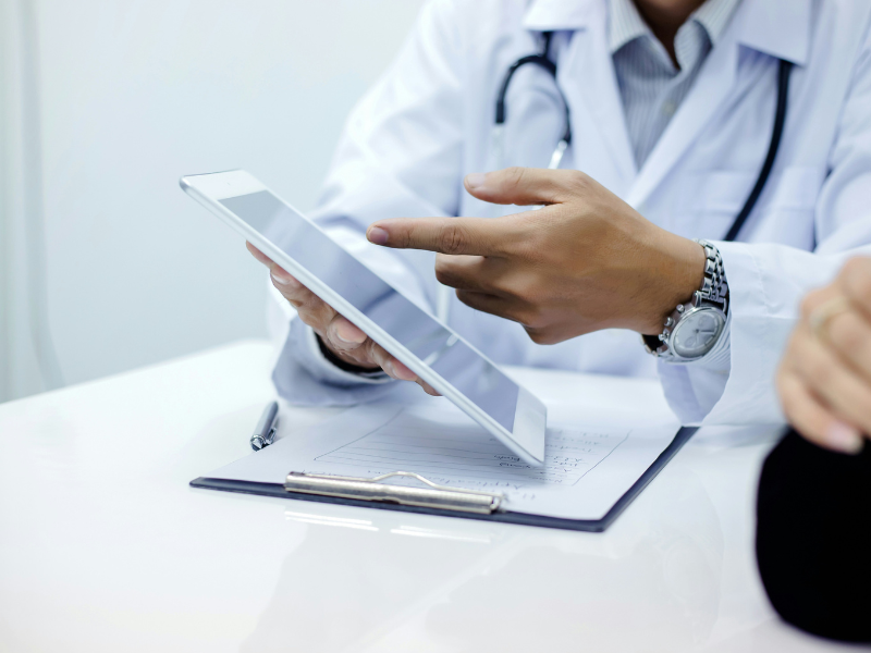 Médico usando tablet em consultório médico, com bloco de notas e caneta na mesa, demonstrando tecnologia na área da saúde.