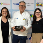 Homem de jaqueta branca segura estojo entre duas mulheres em frente a painel do SUS.